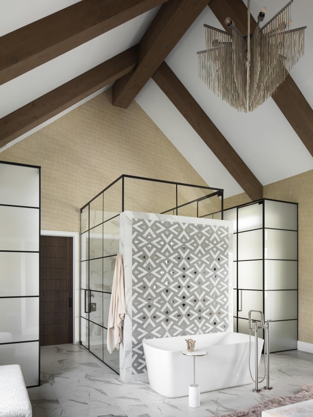 The primary bathroom features a custom tile-and-mirror mosaic by Audrey Lane, which creates a patterned backdrop against Kohler’s pristine white soaking tub. Frosted glass provides privacy for the water closets, while a clear glass shower with dual entrances enhances the room’s ethereal appeal. A metallic leather chandelier from Ngala Trading Co. accentuates the soaring vaulted ceiling. #floridadesign #featured #pinecreek #modernranch #ranchdesign #bathroomdesign